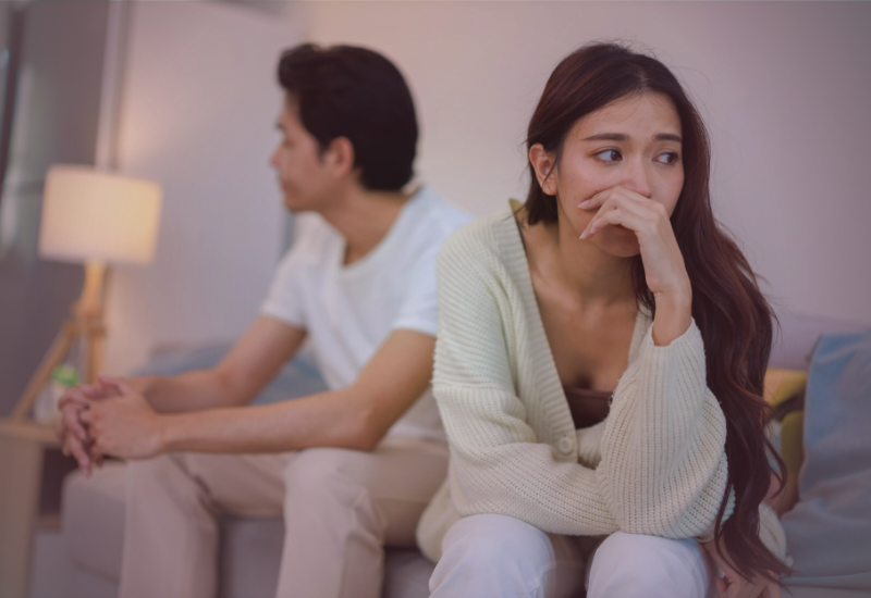 Mulher sentada no sofá, visivelmente triste e pensativa, enquanto o parceiro está ao fundo, afastado emocionalmente, ilustrando o impacto do autoabandono em relacionamentos sem reciprocidade.